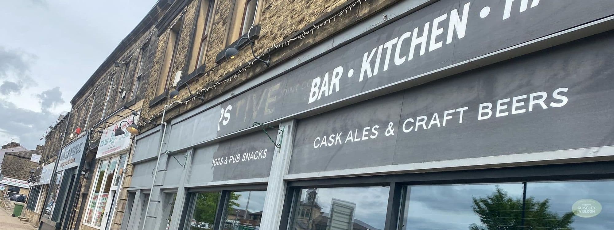 Exterior view of Coopers Bar and Brasserie showcasing its signage with the words "Bar Kitchen" and offerings such as "Cask Ales & Craft Beers." The facade has a dark gray color scheme with large windows and is situated on a street with other storefronts.