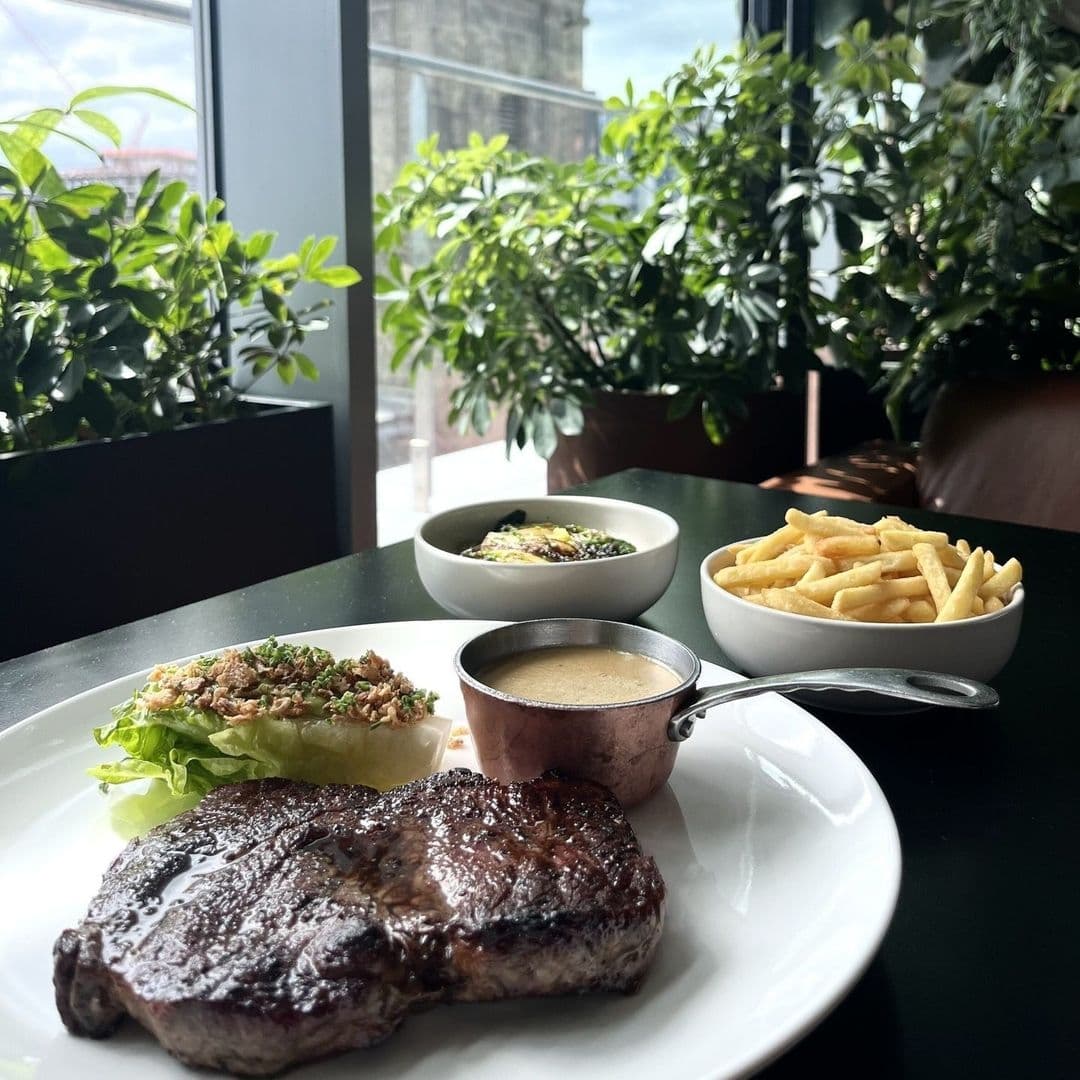 Grilled steak served with a side of fries and sauce at Crafthouse, with a fresh green salad and a bowl of soup, in a dining setting with natural light and green plants.