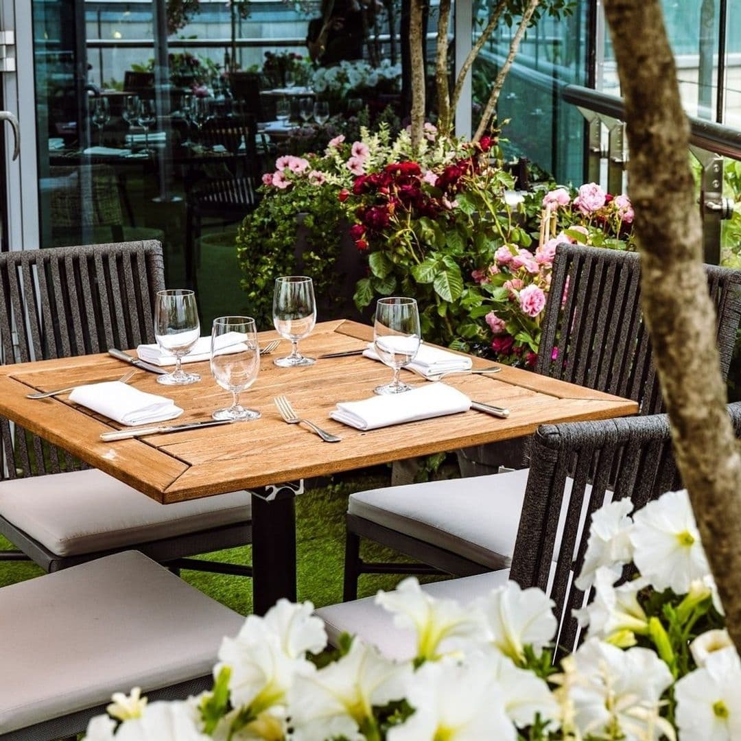 Outdoor dining setup at Crafthouse with a wooden table set for four, surrounded by lush flowers and greenery, with a view of the restaurant's interior through glass windows.