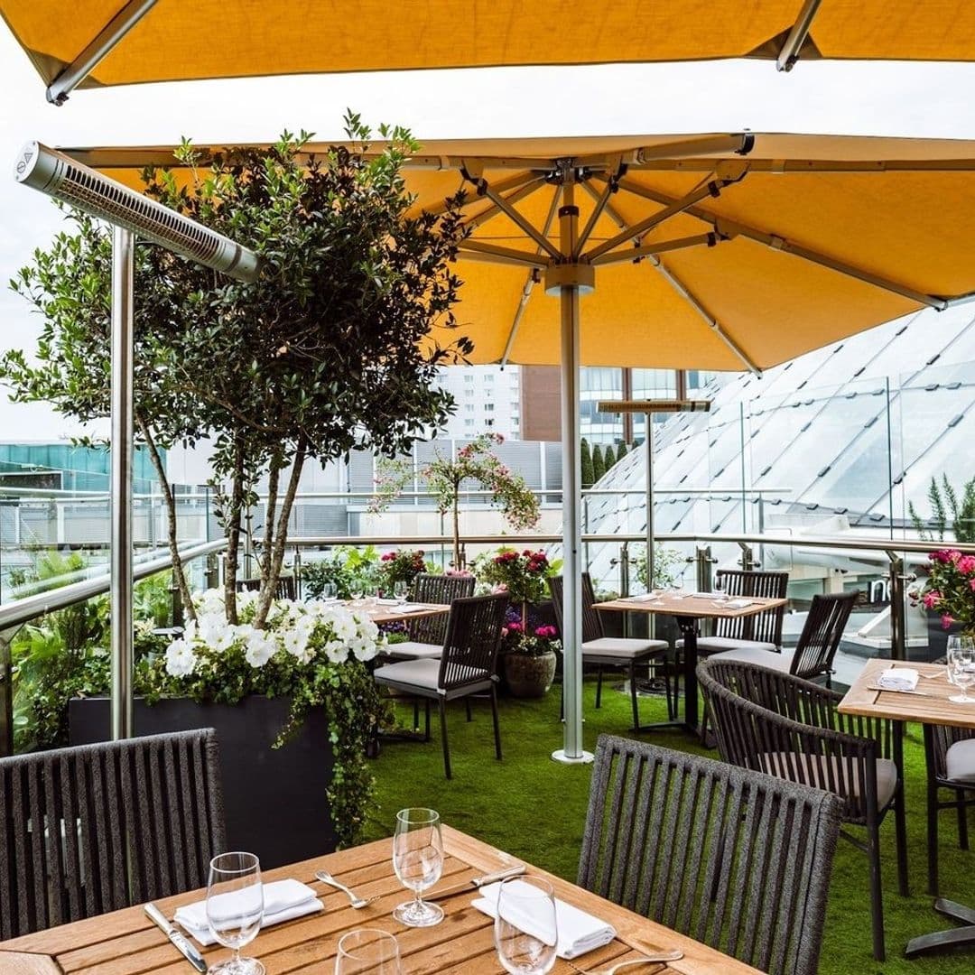 Outdoor dining area at Crafthouse with wooden tables, wicker chairs, and large yellow umbrellas, surrounded by lush greenery and flowers.