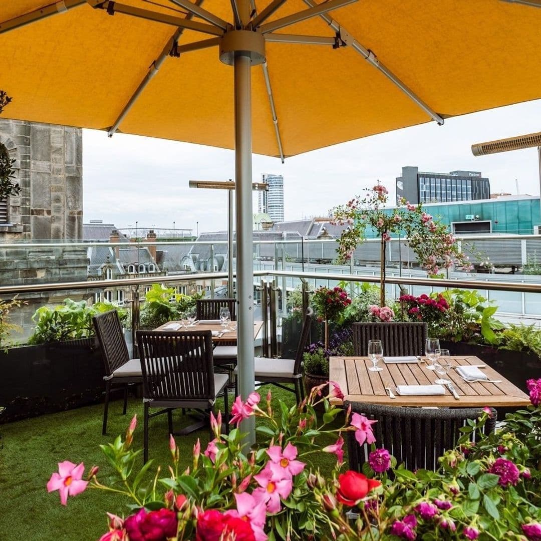 Outdoor dining area at Crafthouse with wooden tables, chairs under a large yellow umbrella, surrounded by vibrant pink and purple flowers, with a view of city buildings in the background.