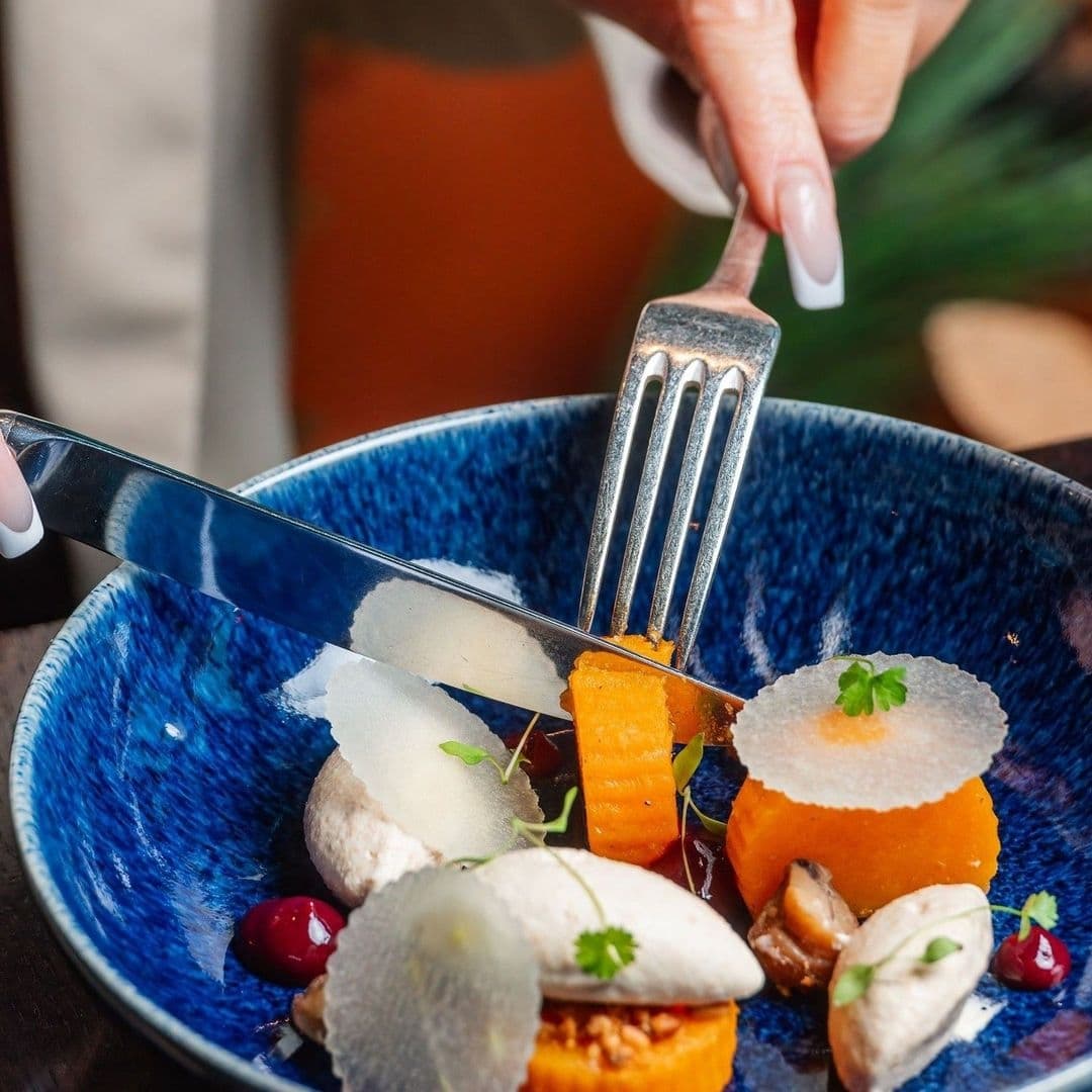 Gourmet dining experience at Crafthouse with a close-up of a hand using a fork and knife to cut into a beautifully plated dish with vibrant orange carrot, delicate garnishes, and artistic presentation in a blue bowl.