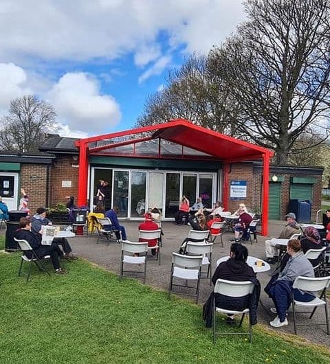 Visitors enjoying outdoor seating at Cross Flatts Park Cafe with a red canopy, surrounded by greenery under a partly cloudy sky.