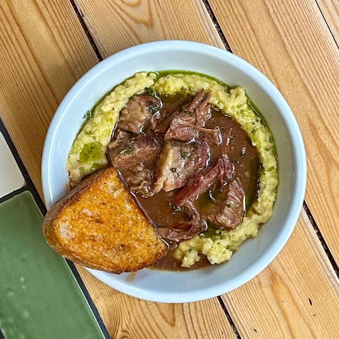 Hearty meal served at Eat Your Greens featuring tender slices of beef on a bed of creamy mashed potatoes with gravy, accompanied by a slice of golden-brown toast, presented in a white bowl on a wooden table.
