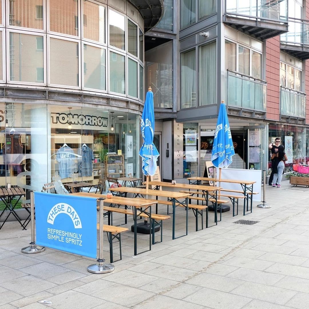 Outdoor seating at Eat Your Greens with wooden benches and blue promotional umbrellas on a city sidewalk.