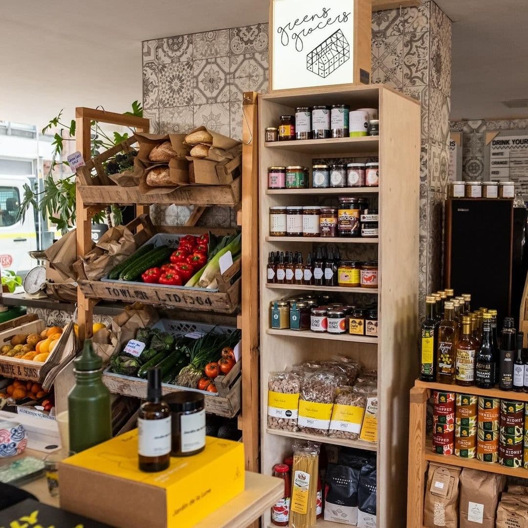 Interior of "Eat Your Greens" store showcasing fresh produce on wooden shelves, artisanal bread, and a variety of packaged organic foods and condiments.