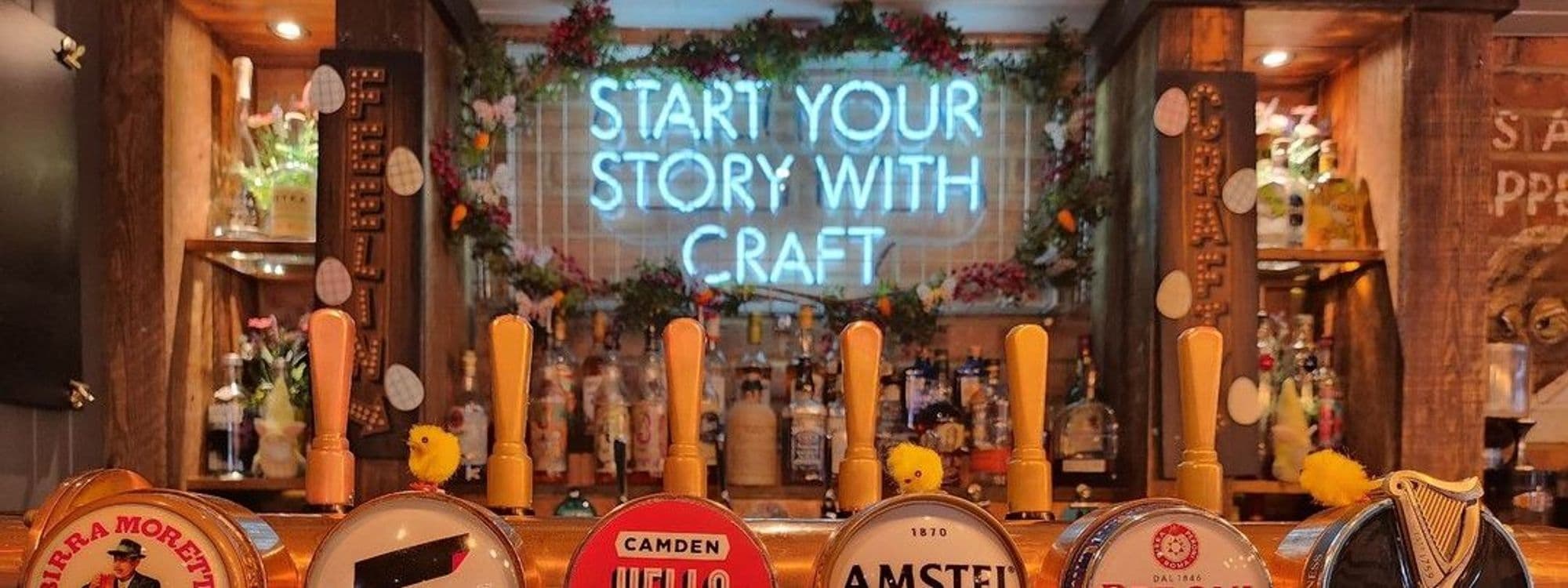 Interior view of Editor’s Draught bar with beer taps in the foreground and a neon sign saying "Start your story with craft" in the background, surrounded by warm lighting and floral decorations.