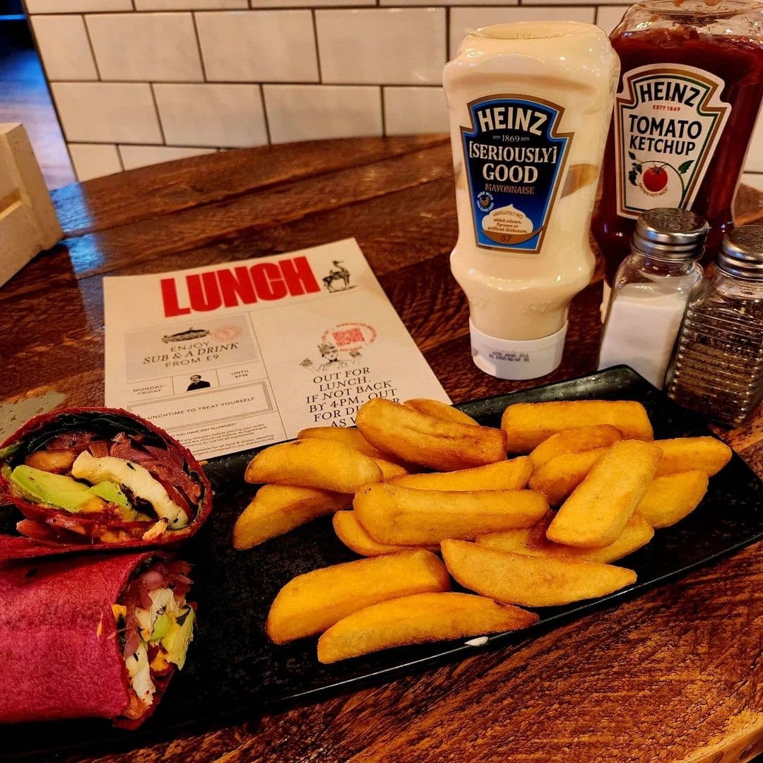 Lunch meal at Editor's Draught featuring a wrap with avocado and chicken, a side of thick-cut fries, Heinz ketchup and mayonnaise bottles, salt shaker, and a menu on a wooden table.