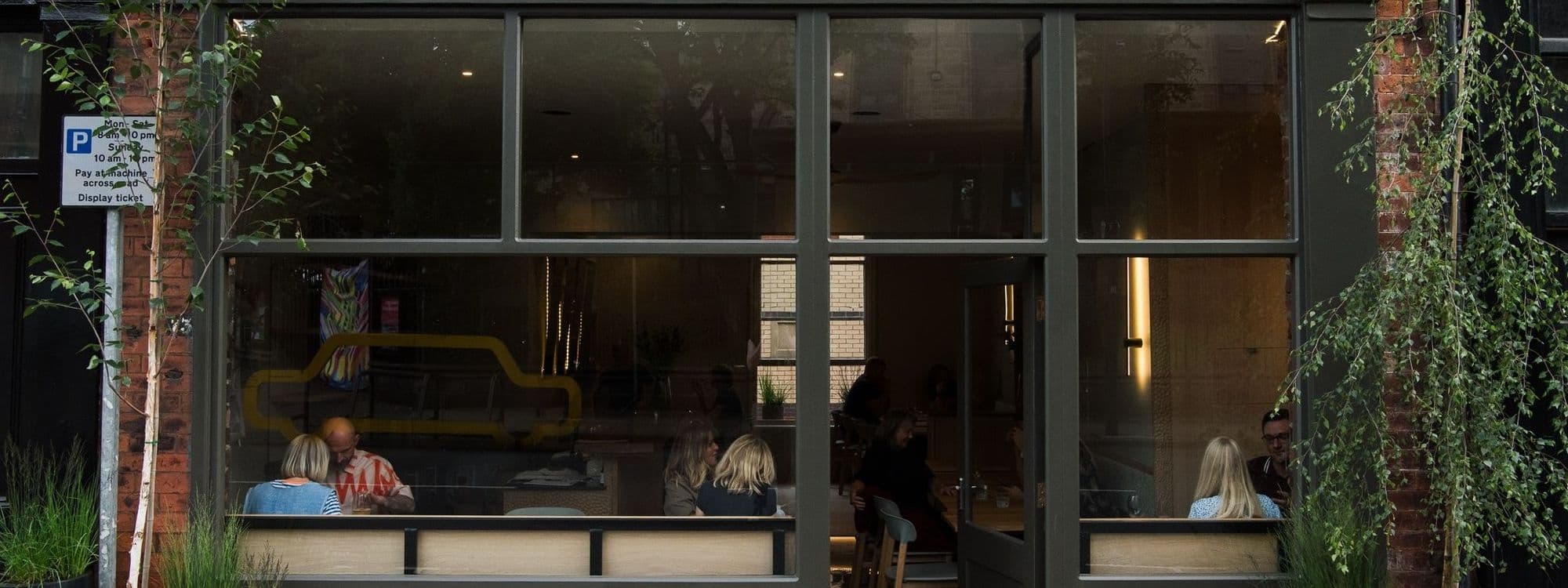 Exterior view of FINT, showing people dining inside through large windows, with greenery and a parking sign visible outside.