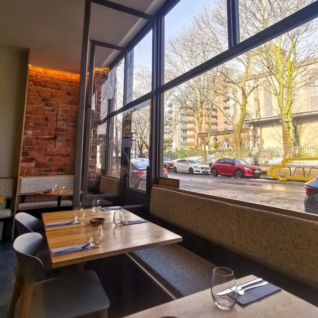 Cozy restaurant interior at FINT with modern decor, exposed brick wall, large windows overlooking a street with trees and parked cars, set tables ready for guests.