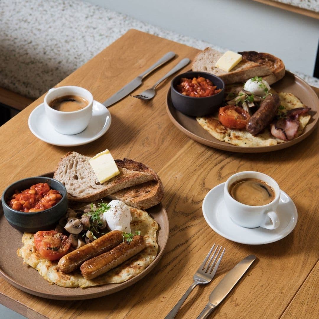 Hearty breakfast at FINT featuring poached eggs, sausages, bacon, grilled tomatoes, beans, mushrooms, toast with butter, and cups of espresso on a wooden table.