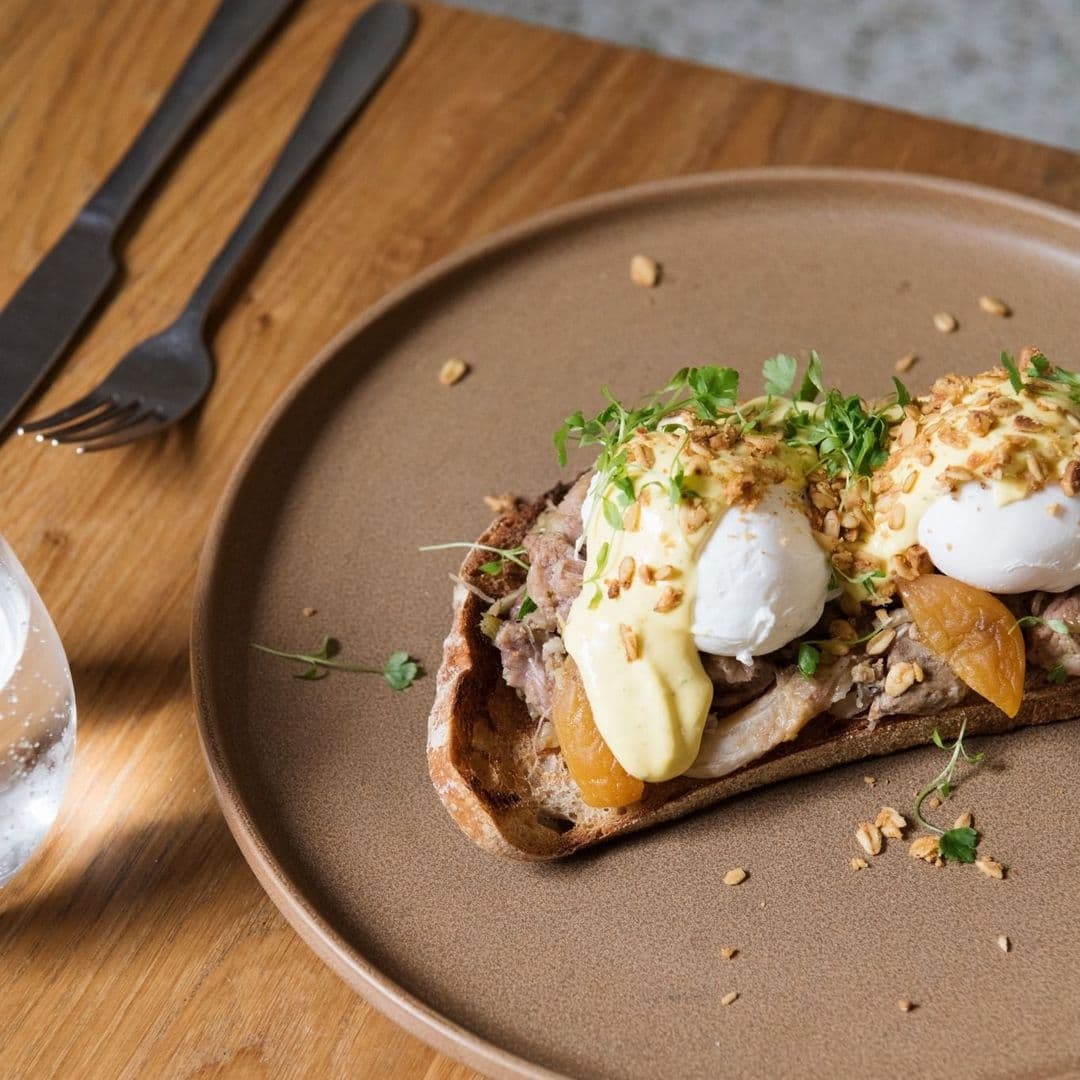 Gourmet open-faced sandwich served on a brown plate at FINT, featuring poached eggs, creamy sauce, nuts, and fresh herbs on artisan bread, with elegant cutlery on a wooden table.