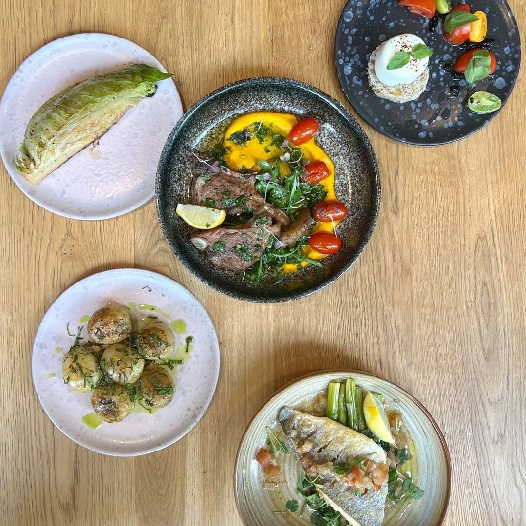Assorted gourmet dishes on a wooden table at FINT, featuring grilled fish with lemon, herb-seasoned potatoes, a salad with creamy dressing, and a steak with yellow sauce and tomatoes.