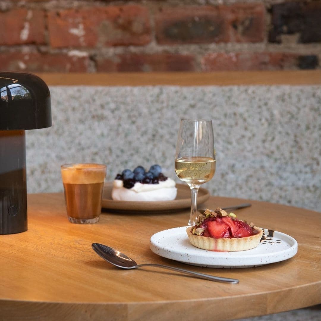 Alt text: "Delicious dessert and drinks at FINT, featuring a fresh fruit tart with strawberries and nuts on a white plate, a glass of white wine, and a cup of espresso on a wooden table with a rustic brick wall in the background."