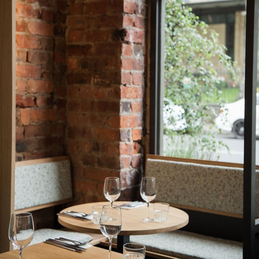 Cozy dining setup at FINT with wine glasses on a wooden table, exposed brick wall, and a view of greenery outside the window.