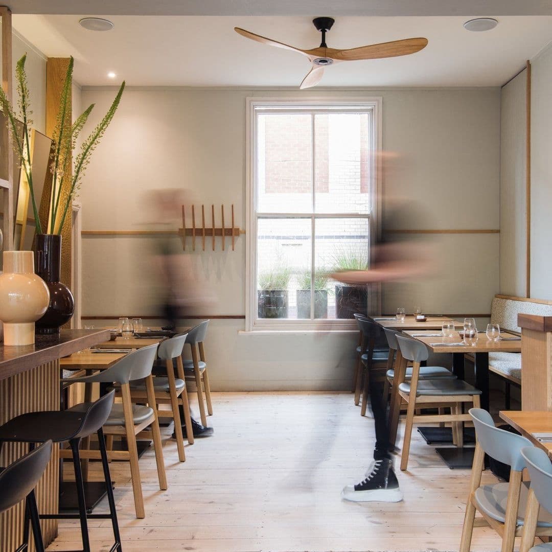 Interior of FINT restaurant with modern minimalist design, featuring wooden tables and chairs, a ceiling fan, and blurred figures of people moving.