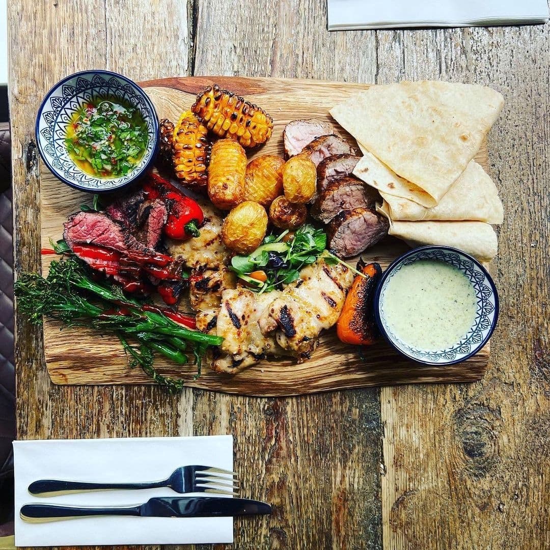 Alt text: "A delicious platter of grilled meats, including steak and chicken, with sides of roasted corn, potatoes, and flatbread, served on a rustic wooden board at a dining venue in Forde."