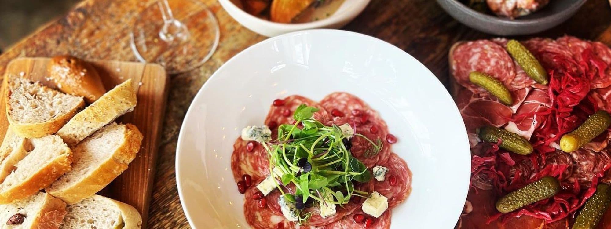 Gourmet dining experience in Forde featuring a platter of sliced bread, a bowl of salami garnished with pomegranate and blue cheese, and a plate of cured meats with pickles and red cabbage.