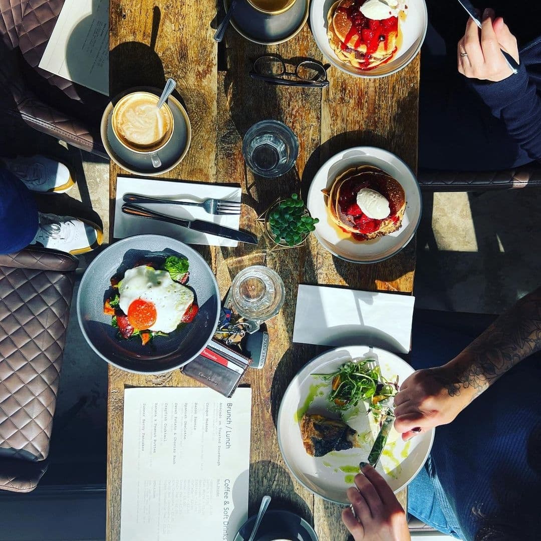 Alt text: "A cozy dining experience in Forde featuring a sunny table with a variety of breakfast dishes, including pancakes with syrup and a fried egg on toast, accompanied by a cappuccino and fresh water, with diners enjoying their meal."