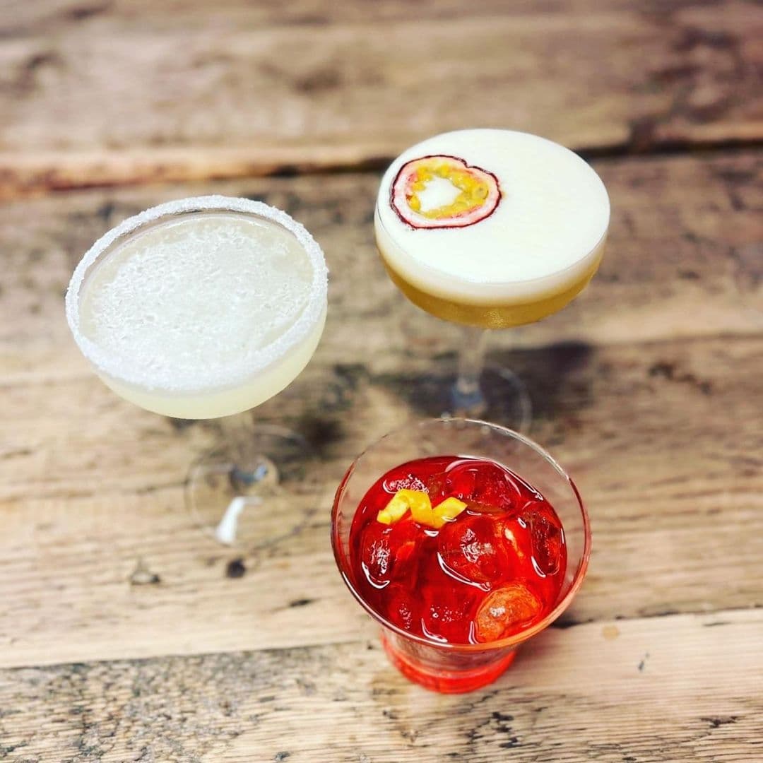 Three colorful cocktails on a wooden table at a Forde bar, featuring a salt-rimmed margarita, a frothy pisco sour garnished with a slice of star fruit, and a vibrant red negroni with an orange peel twist.