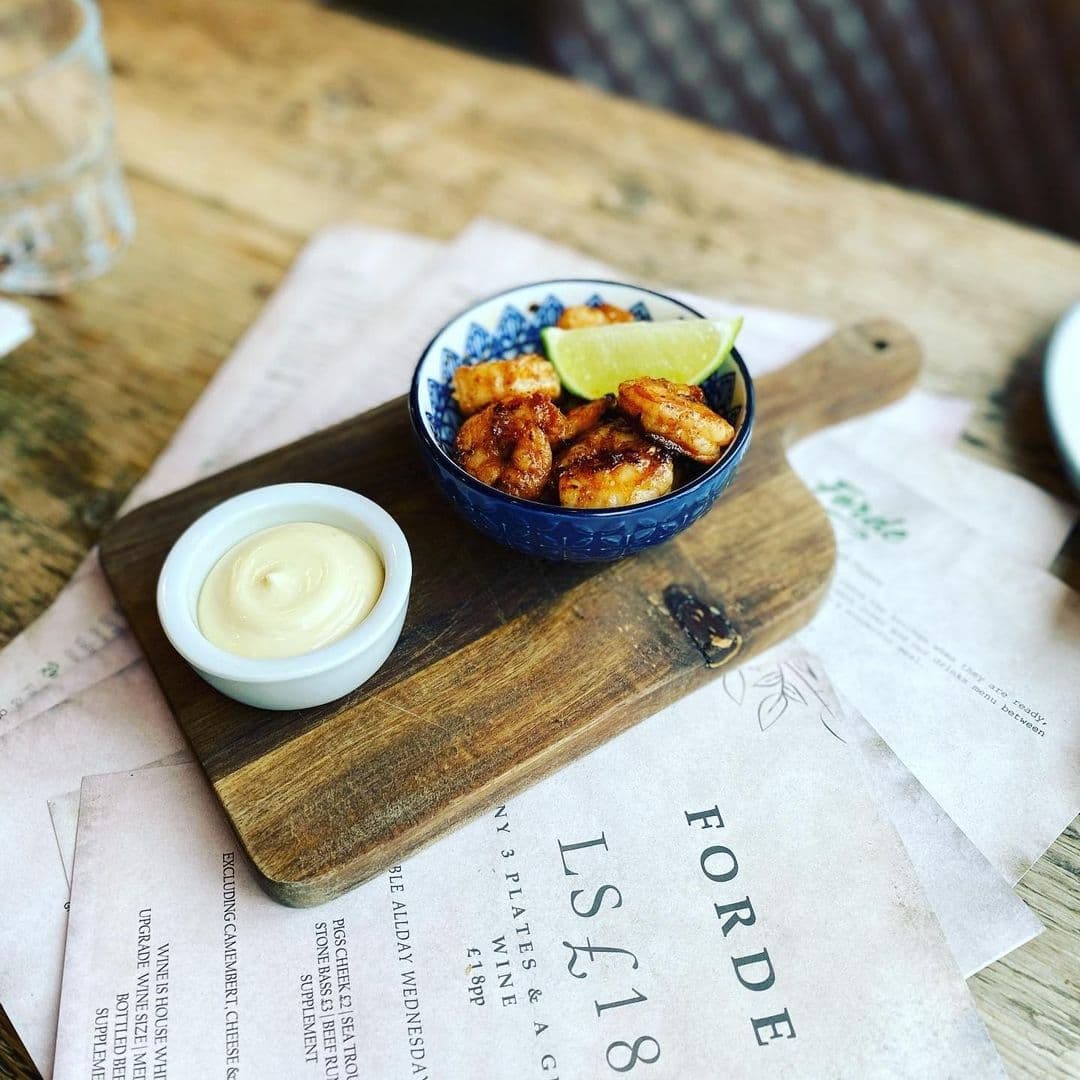 Appetizing dish served at Forde, featuring grilled shrimp with a lime wedge in a blue patterned bowl, accompanied by a side of creamy sauce, presented on a wooden board with a menu underneath.