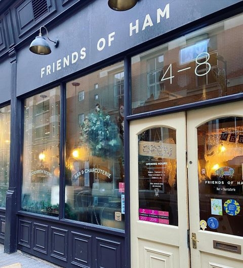 Exterior view of Friends of Ham in Leeds City, showcasing the entrance with a beige door and large windows, the name of the establishment in large white letters, and a sign indicating it is a bar and charcuterie.