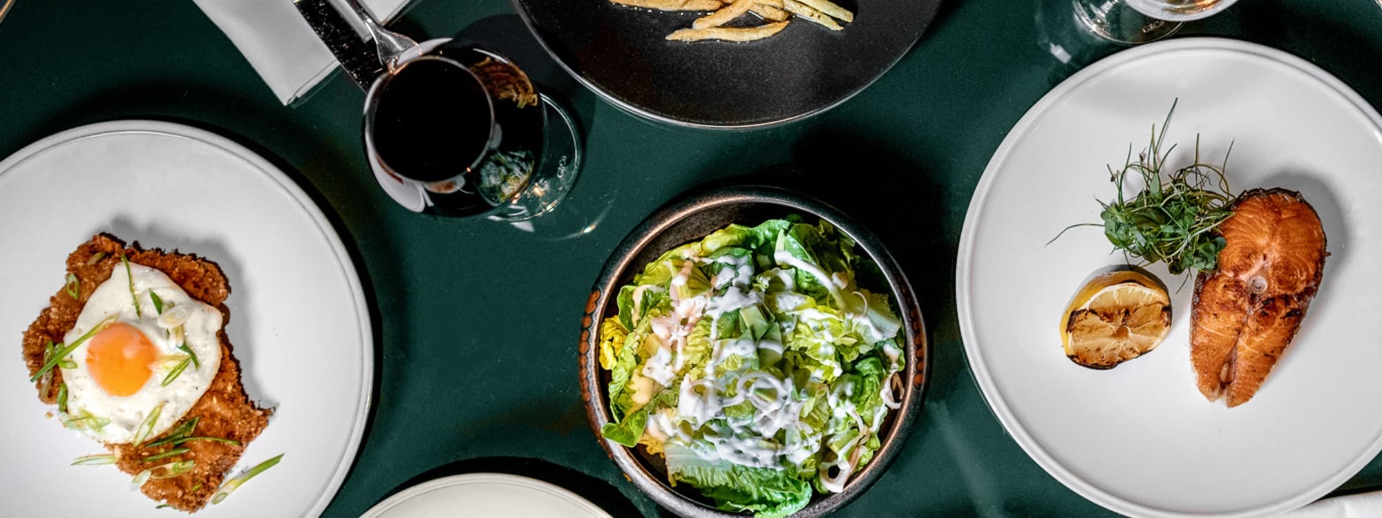 Overhead view of a dining table at Gaucho featuring a variety of dishes including a breaded cutlet topped with a fried egg, a fresh green salad with dressing, and a grilled fish with lemon, accompanied by a glass of red wine.