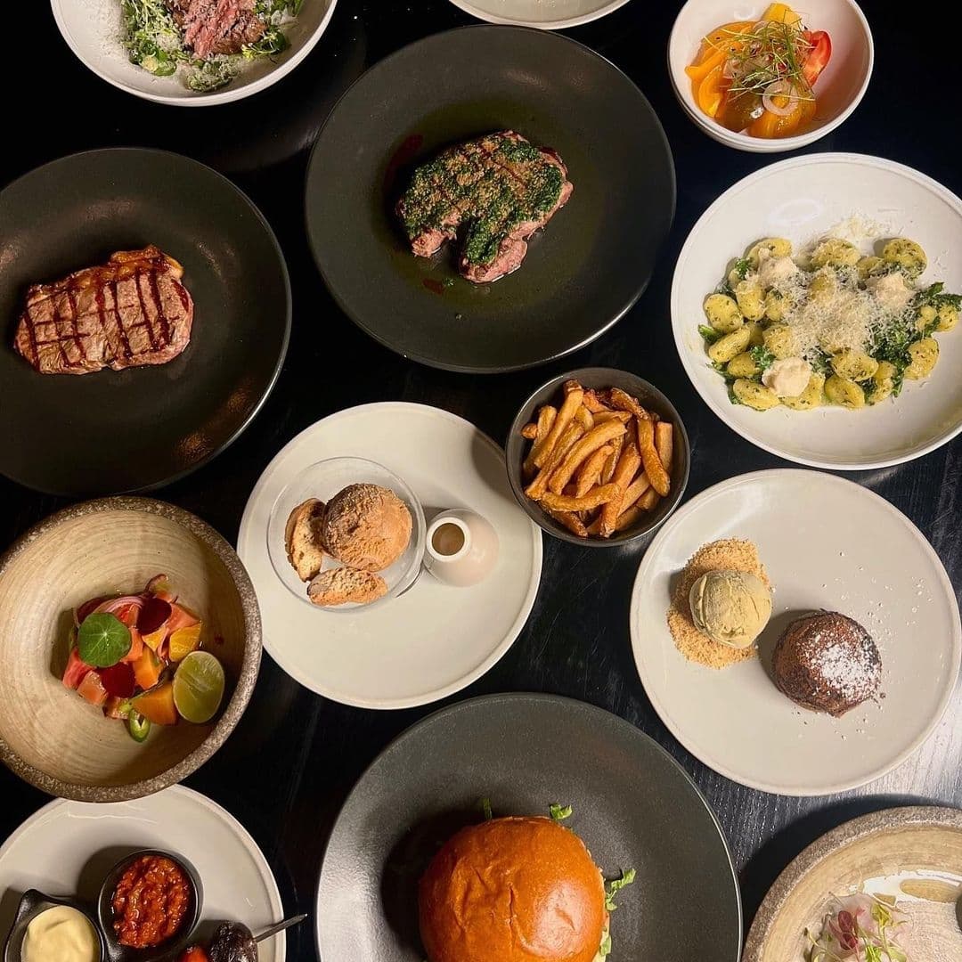 Assorted dishes at Gaucho restaurant featuring grilled steak, pasta, fries, bread, and desserts on a dark table.