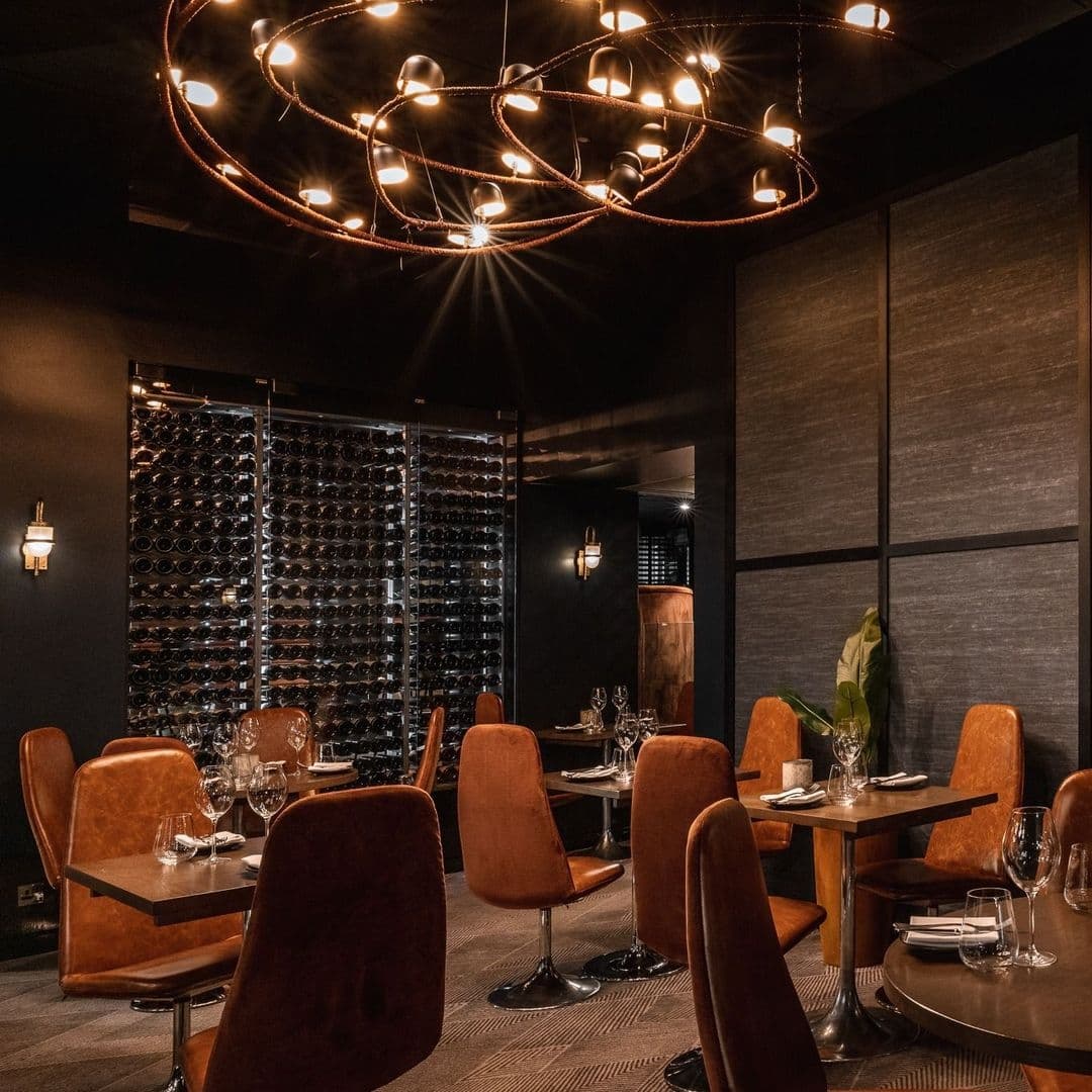 Elegant dining area at Gaucho with plush velvet chairs, modern circular lighting fixture, and a wall of wine bottles.