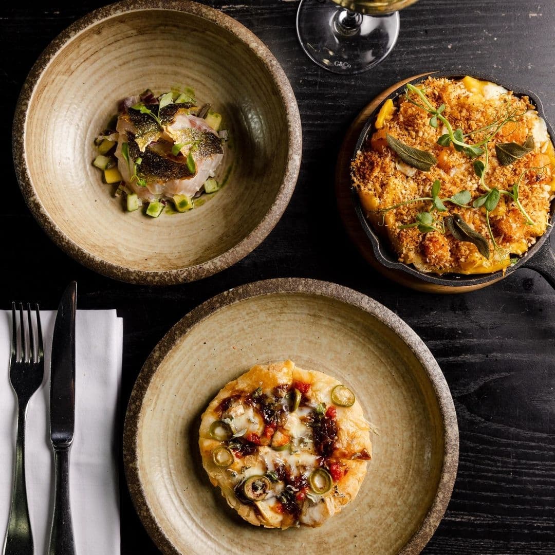 Alt text: "Gourmet dishes served at Gaucho, featuring a bowl of ceviche, a macaroni cheese gratin with sage, and a pizza with olives and chili oil, all presented on rustic ceramic plates against a dark wooden table."