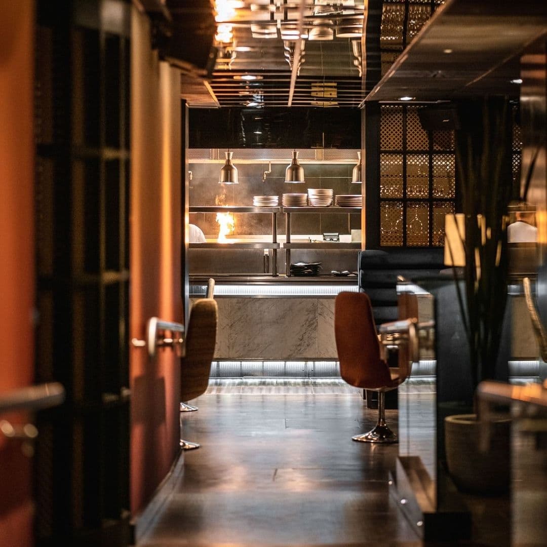 Interior view of Gaucho restaurant showcasing the open kitchen with a visible flame, warm ambient lighting, and modern furnishings.