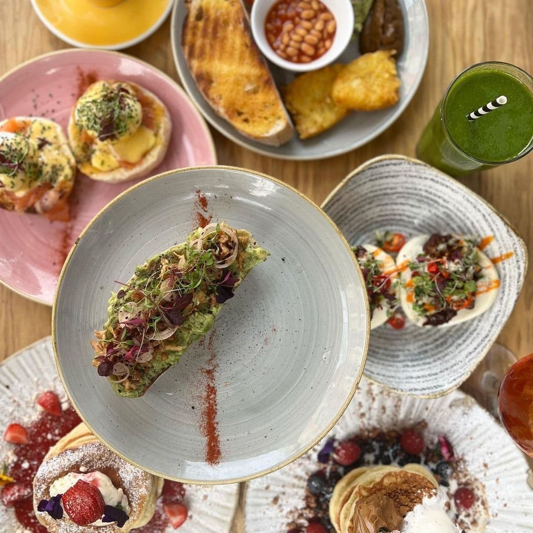 Assorted brunch dishes and drinks on a wooden table at IF Bar – Call Lane, featuring avocado toast, eggs benedict, fresh pastries with berries, and a green smoothie.
