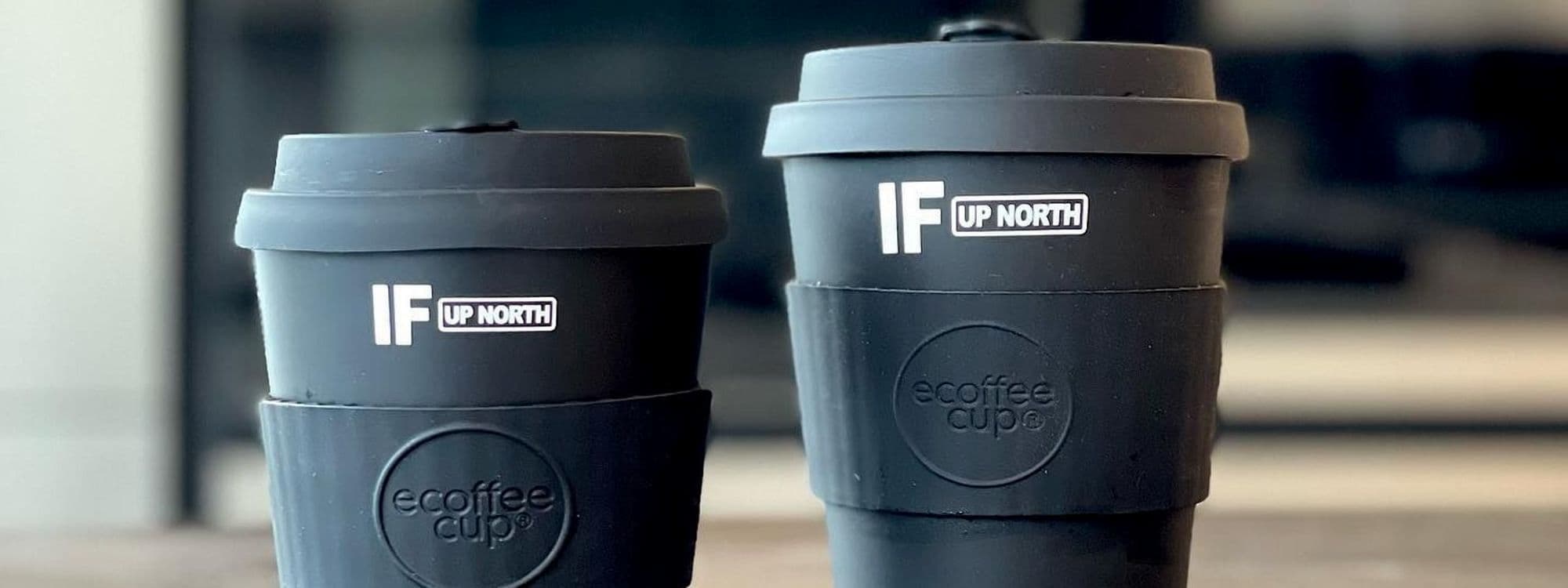 Two reusable black coffee cups with 'IF UP NORTH' branding on a table, representing a sustainable option for visitors at IF Bar – Call Lane.