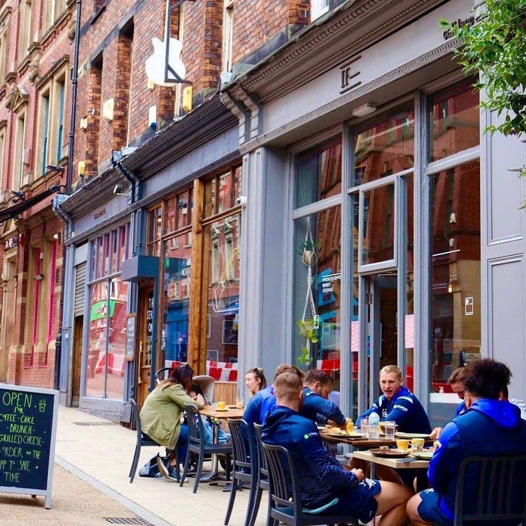 Exterior view of IF Bar – Call Lane with patrons dining at outdoor tables on a city sidewalk, featuring a grey facade and large windows, adjacent to traditional brick buildings.