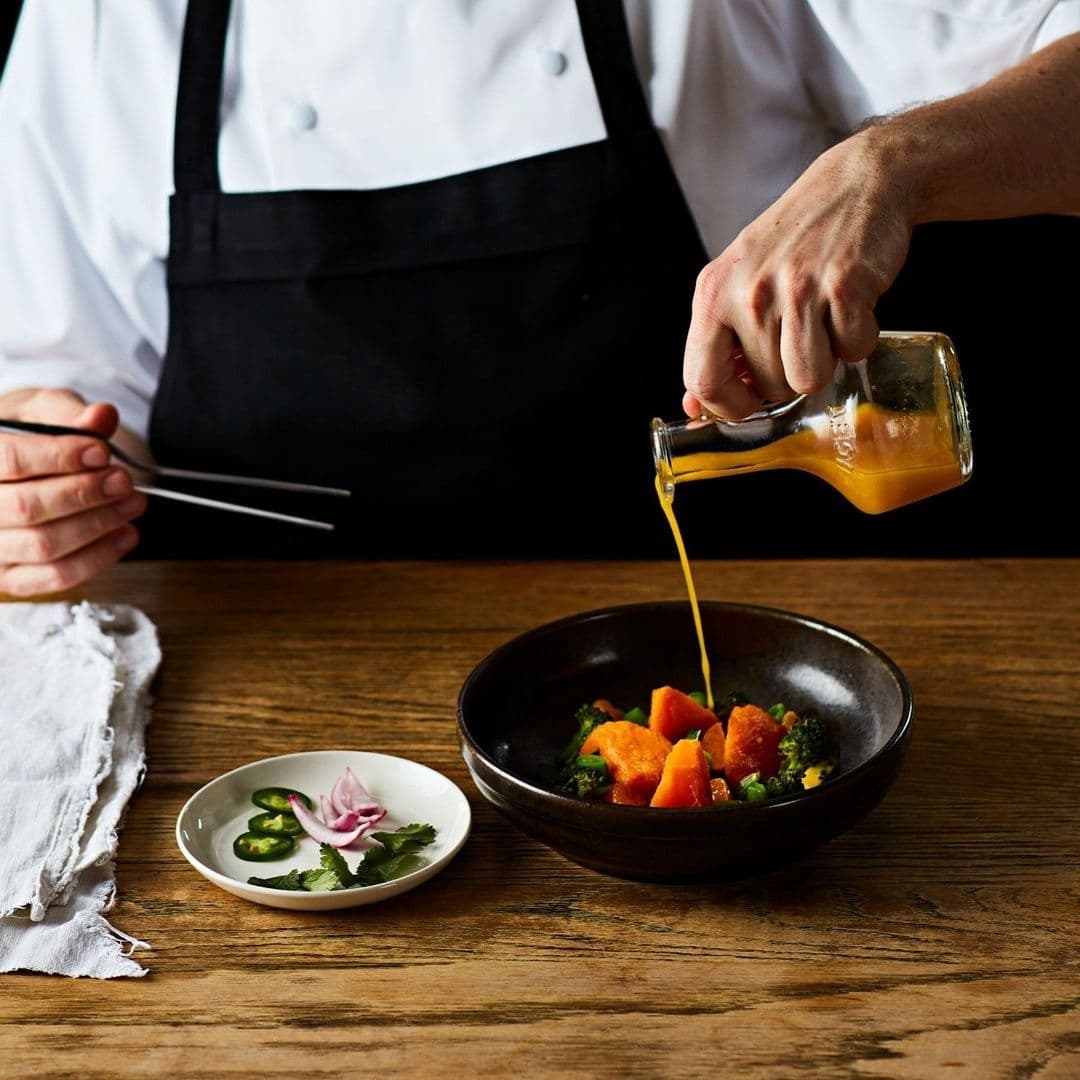 Chef in a white uniform with a black apron pouring sauce over a colorful vegetable dish at Ibérica.