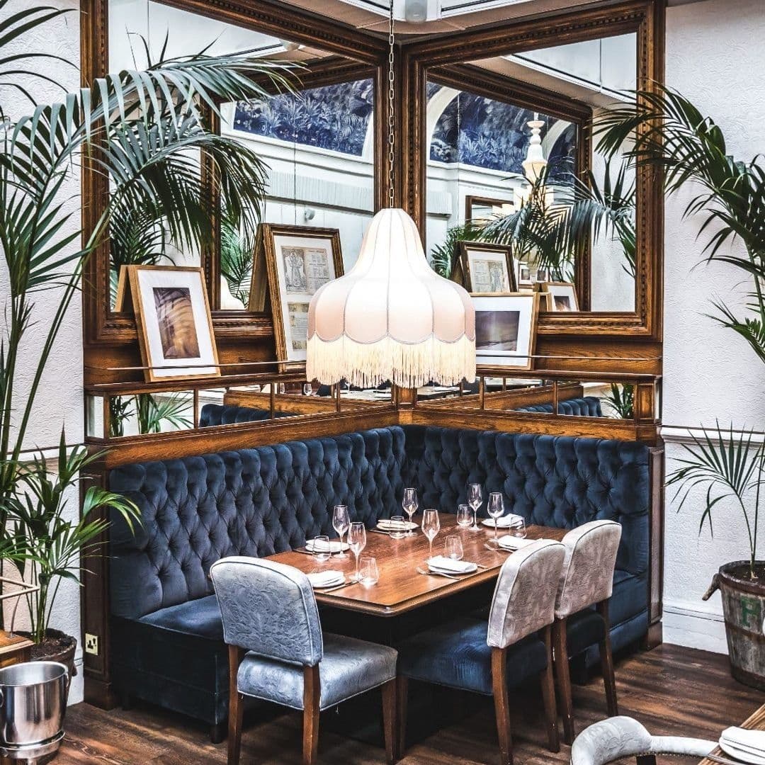 Elegant dining area at Ibérica with blue tufted booth seating, a classic wooden table set for a meal, assorted framed pictures on the wall, a large white pendant lamp with fringe, and potted green plants.