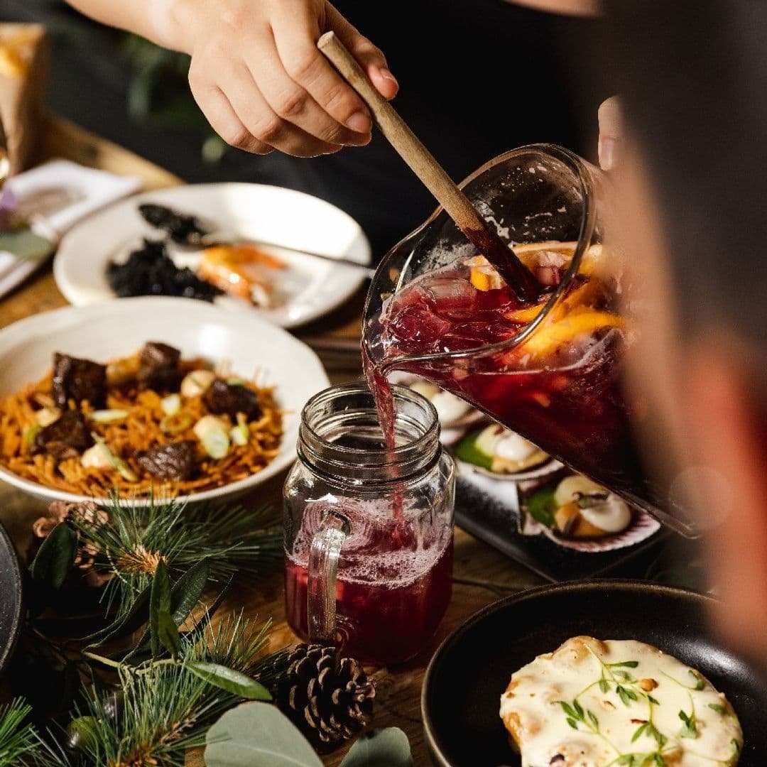 Alt text: "A person pouring sangria from a glass pitcher into a mason jar at Ibérica, surrounded by an assortment of Spanish dishes including paella and tapas, evoking a warm and inviting dining experience."