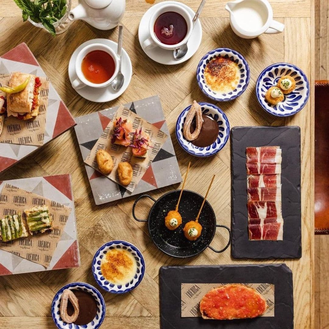 Assorted Spanish tapas and desserts on a wooden table at Ibérica, featuring sandwiches, croquettes, Iberian ham, crème brûlée, and cups of tea.