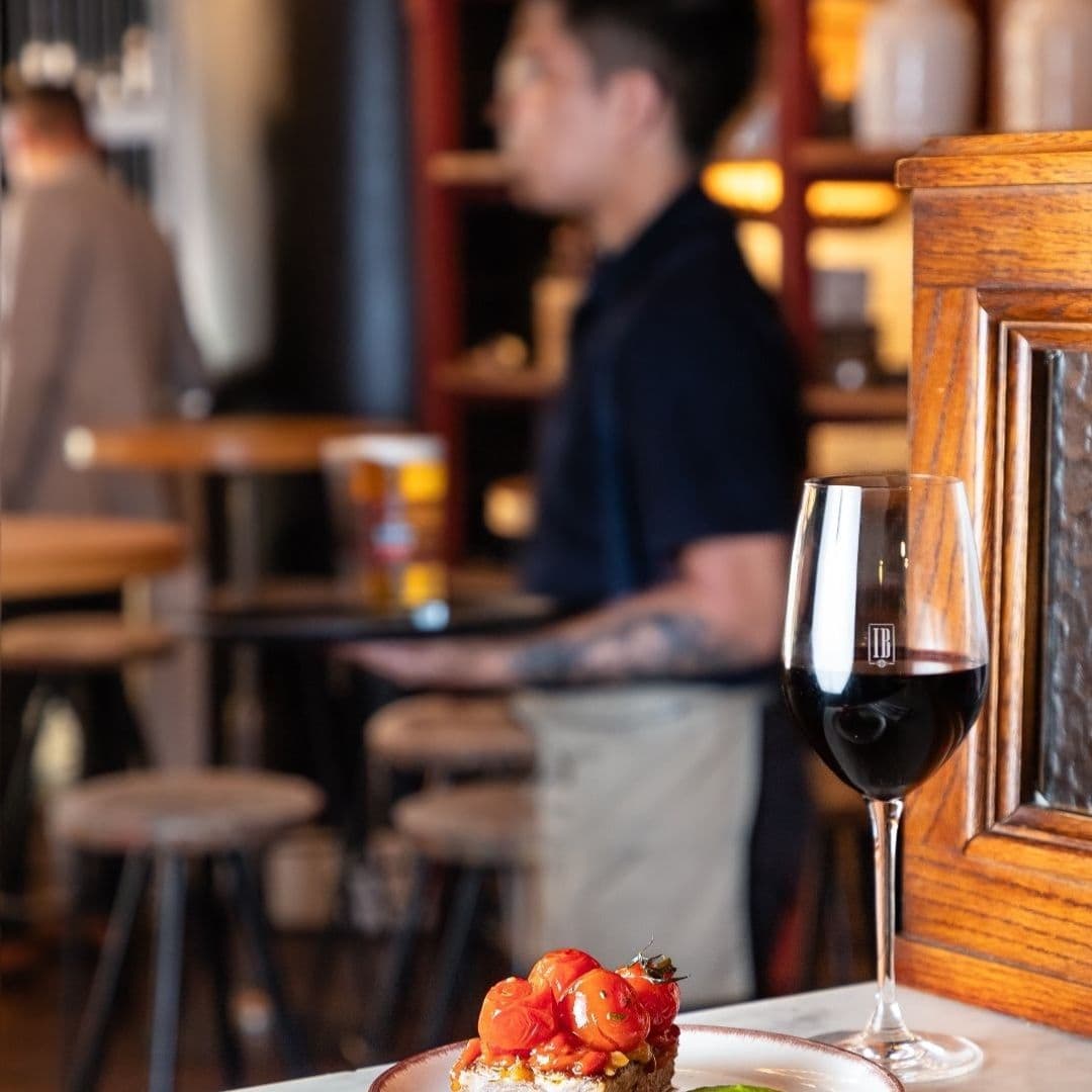 Alt text: "Cozy ambiance at Ibérica with a close-up of a glass of red wine and a plate of tapas topped with cherry tomatoes, with a blurred background of a waiter and customers."