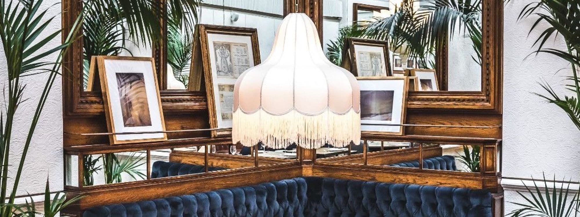Elegant interior of Ibérica featuring a classic chandelier with fringe detail, framed artwork on wooden shelves, and plush blue seating surrounded by lush green plants.