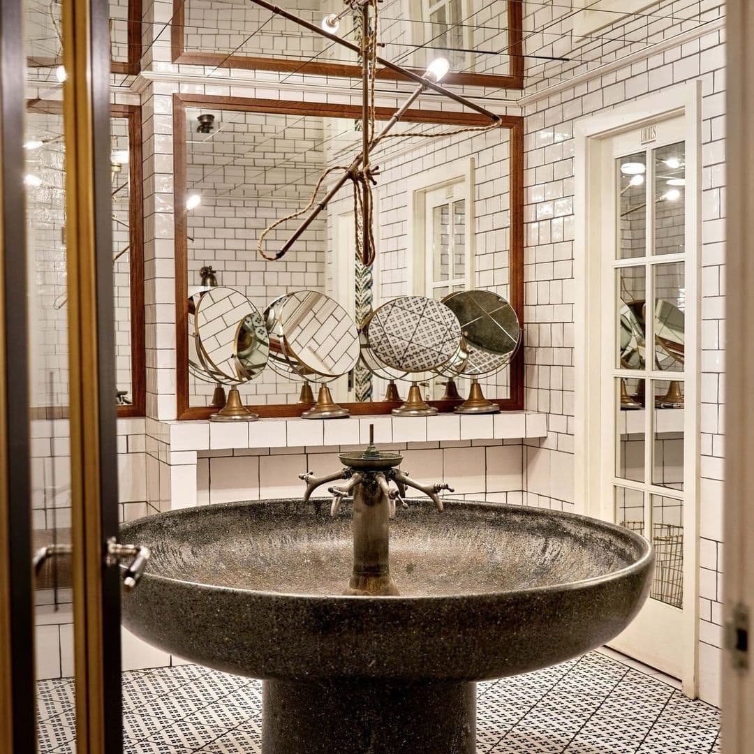 Vintage-style bathroom at Ibérica with a large stone sink, brass faucets, and a mirror surrounded by smaller decorative mirrors on a white subway-tiled wall.
