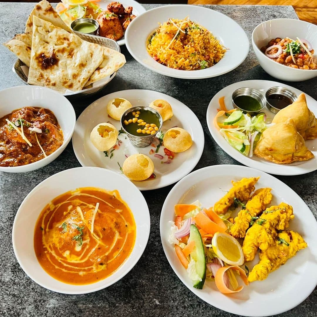 Assorted traditional Indian dishes from Indian Pakwaan, including butter chicken, dal, naan bread, biryani, samosas, pani puri, and fried fish, served on white plates on a grey tabletop.