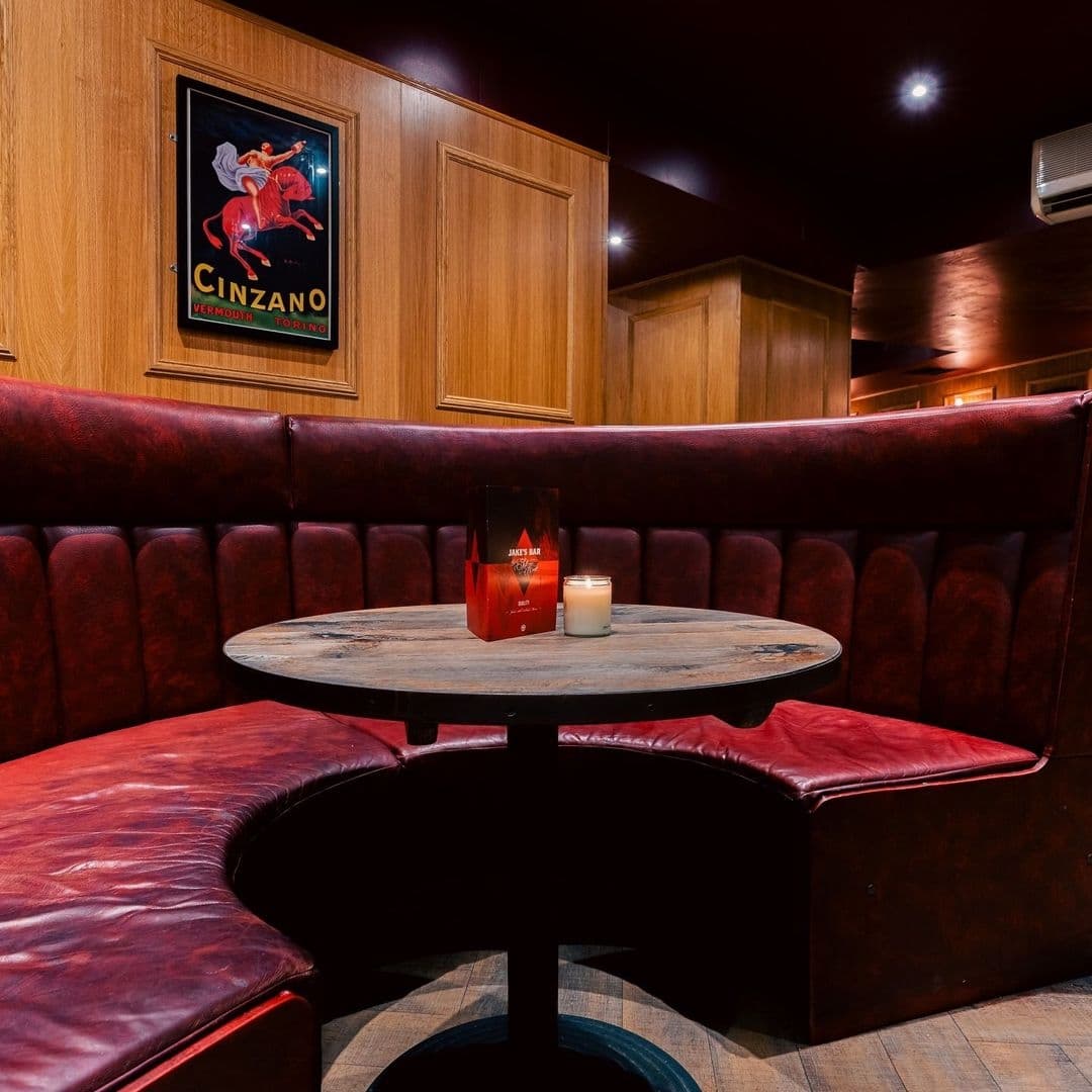 Cozy corner booth with red upholstery at Jake's Bar & Still Room, featuring a wooden round table with a lit candle and a menu stand, under warm lighting with a vintage Cinzano poster on the wood-paneled wall.