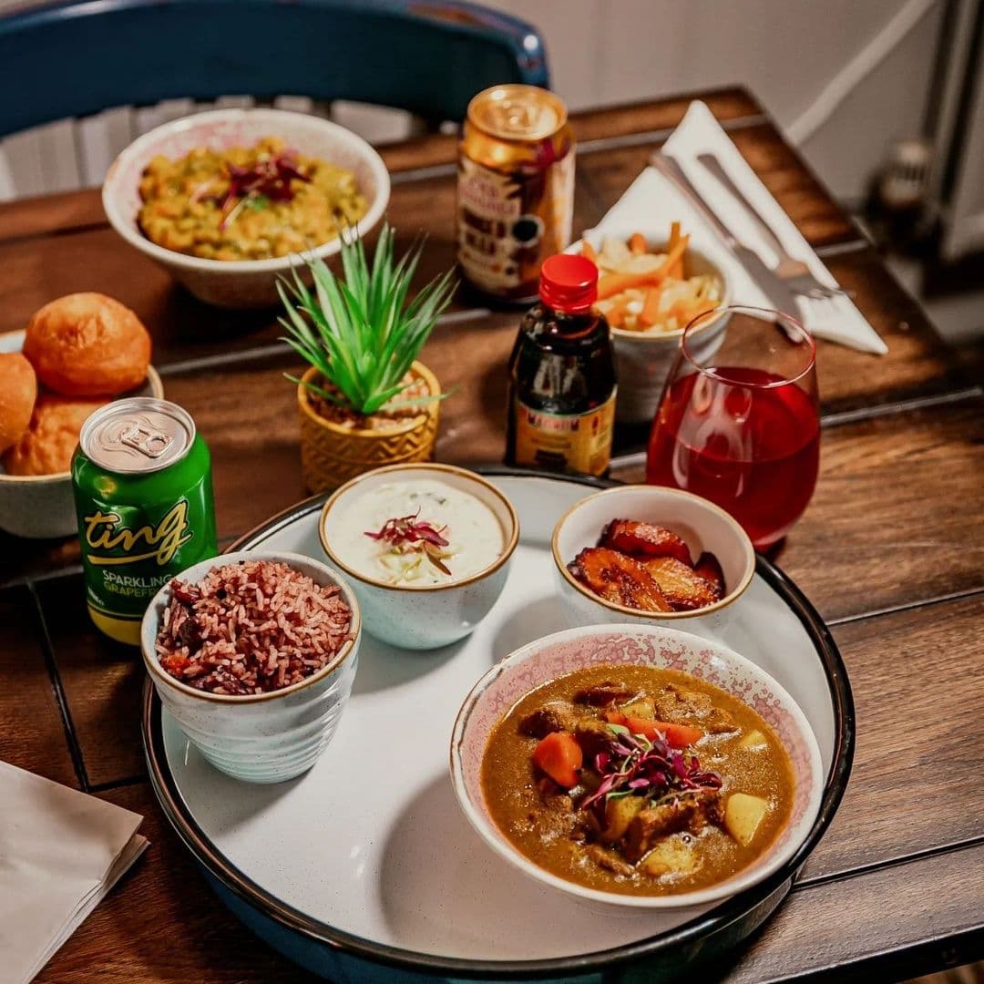 Assorted traditional Jamaican dishes and beverages served on a wooden table at Jamrock, featuring rice and peas, a bowl of soup, fried plantains, a can of Ting soda, and a red drink in a glass.