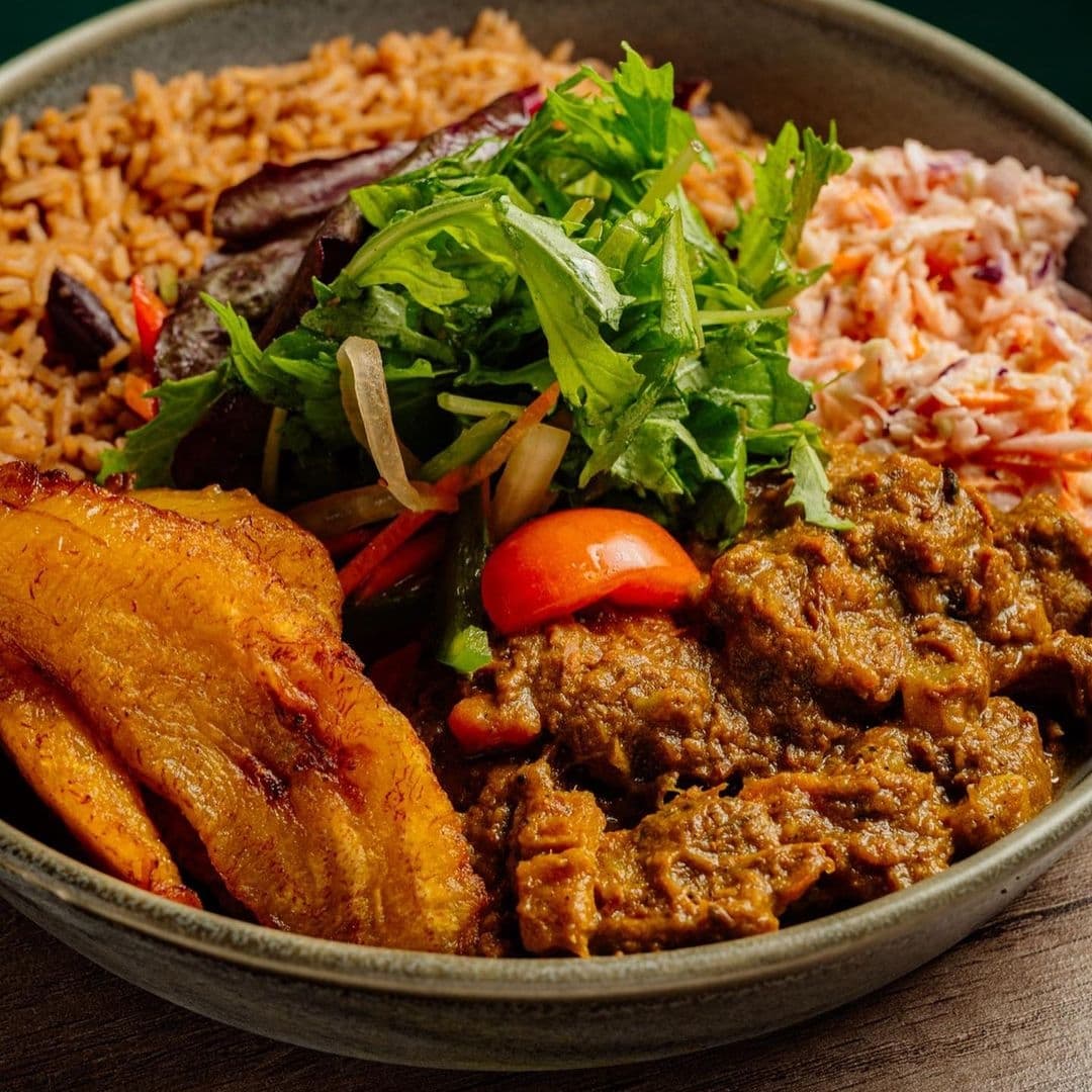 Traditional Jamaican cuisine at Jamrock featuring jerk chicken, rice and peas, fried plantains, coleslaw, and fresh salad in a bowl.