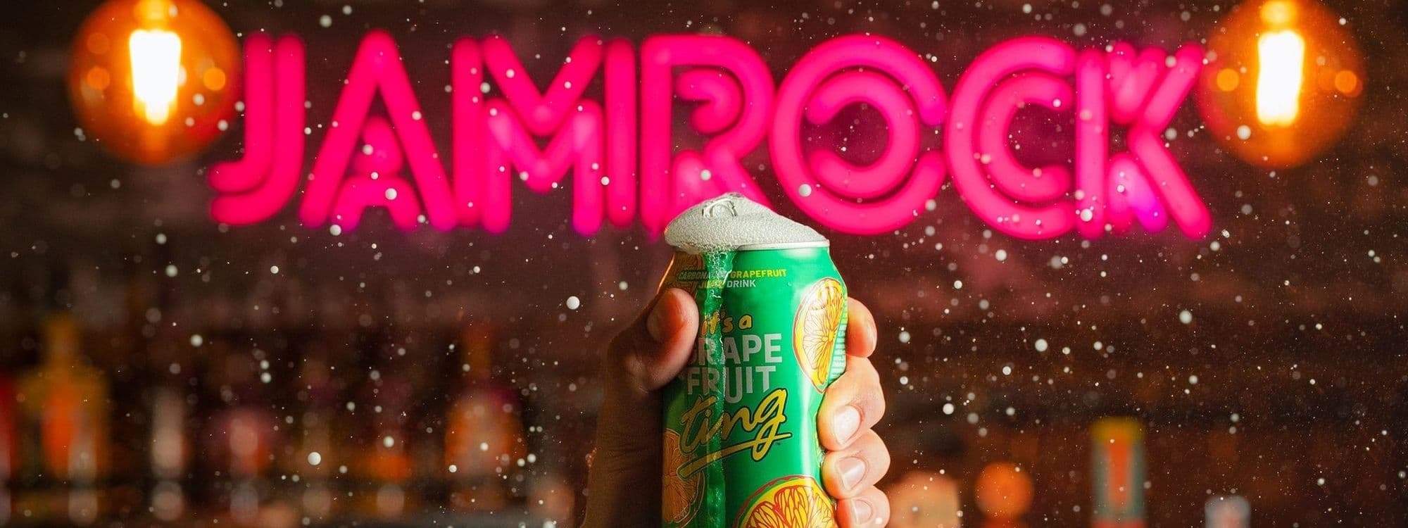 Hand holding a can of grapefruit drink with neon sign reading "JAMROCK" in the background.