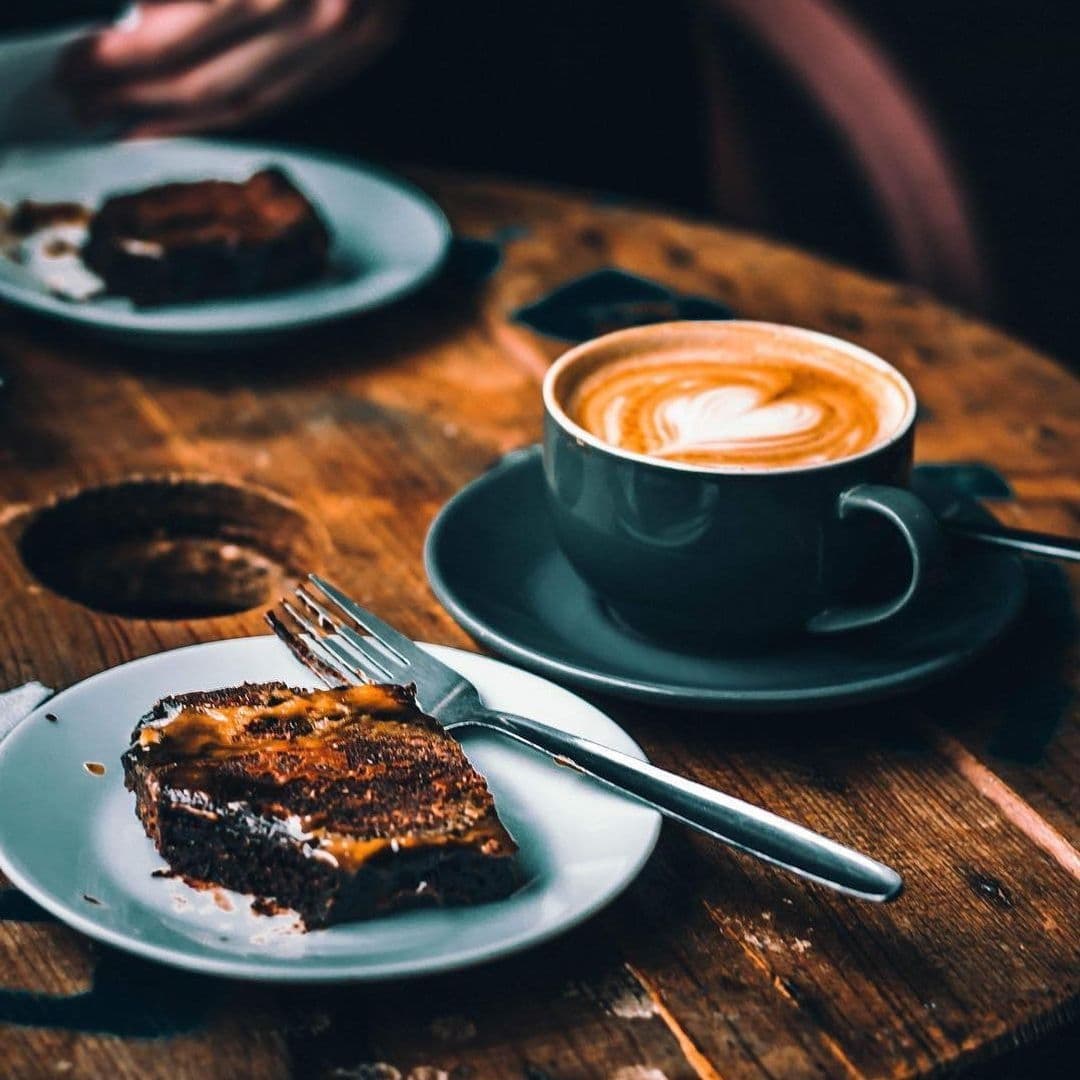 Alt text: "A cozy setting at Kapow Coffee – The Calls featuring a freshly brewed cup of cappuccino with artful foam next to a slice of chocolate cake on a rustic wooden table."
