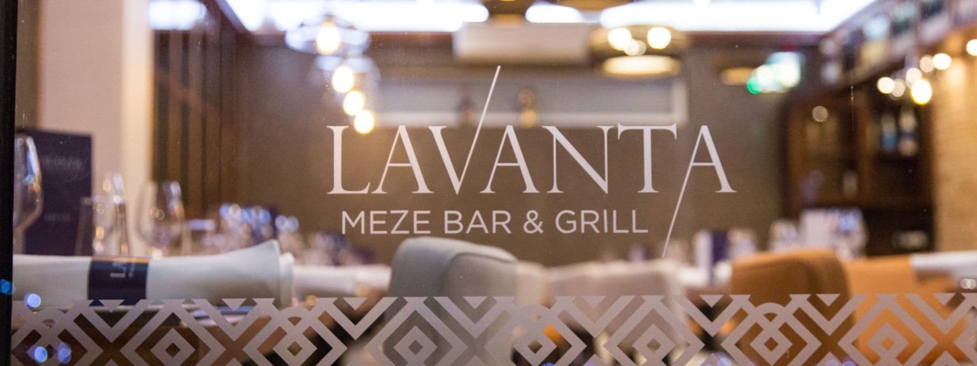Interior view of Lavanta Meze Bar & Grill, showcasing the restaurant's name on the glass window with a decorative frosted pattern, and a blurred background featuring set tables with glassware and warm lighting.
