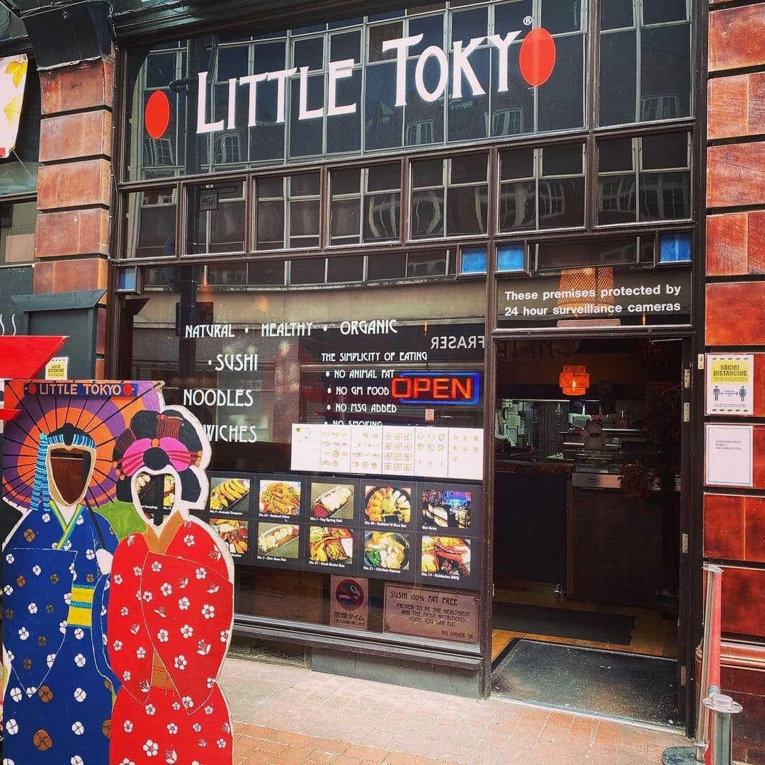 Exterior view of Little Tokyo restaurant with signage displaying 'Natural Healthy Organic' food options such as sushi and noodles, and two colorful traditional Japanese-style cutouts of figures in kimonos at the entrance.