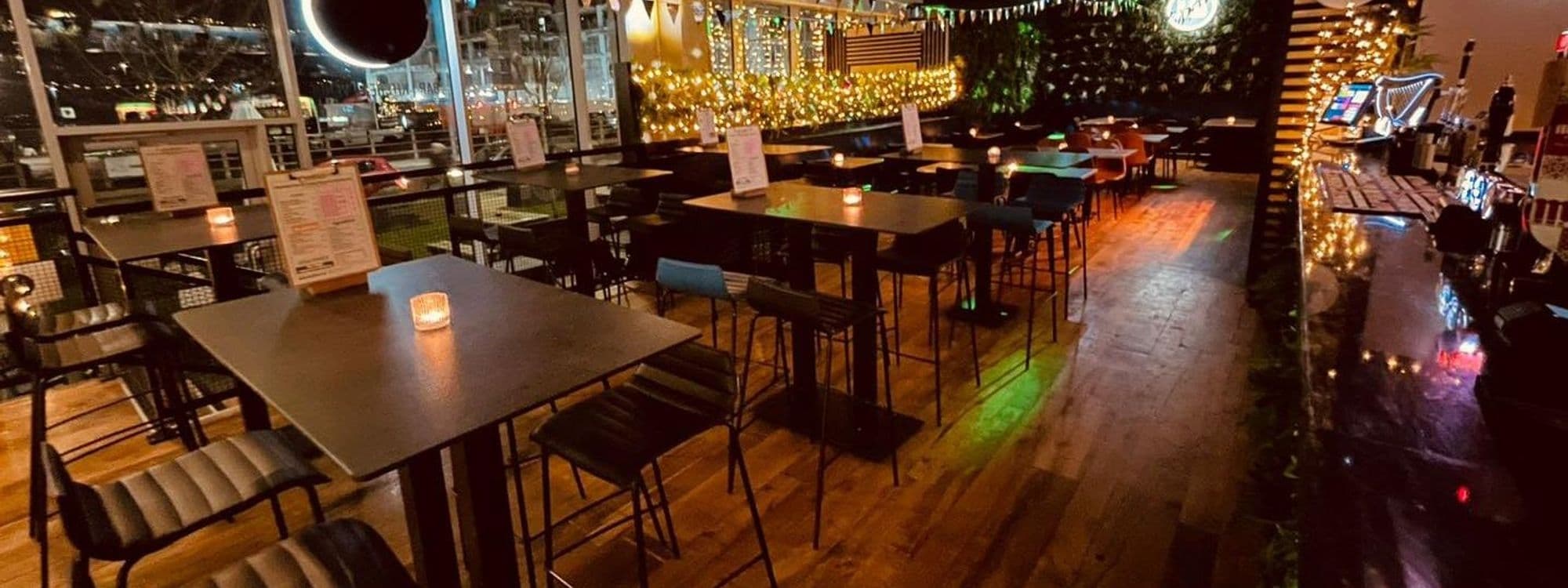 Interior of Mad Frans Bar with wooden tables and chairs, festive string lights, and a well-stocked bar counter illuminated by warm ambient lighting.